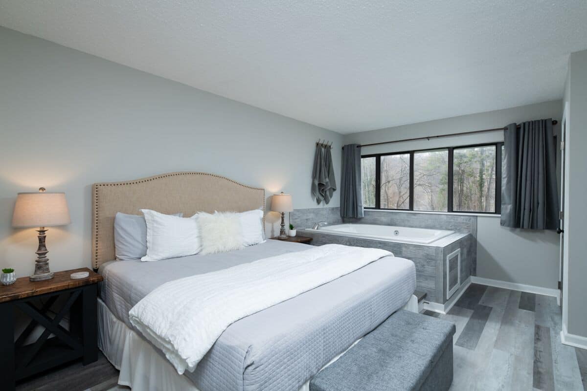 A cozy bedroom featuring a large bed with white linens, a wooden nightstand with a lamp, and a modern bathtub beside a window with curtains.