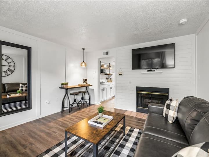 Cozy living room featuring a dark leather couch, a coffee table with decor, a flat-screen TV mounted on the wall, and a modern-style mirror. Wooden flooring complements the warm lighting and stylish furnishings.