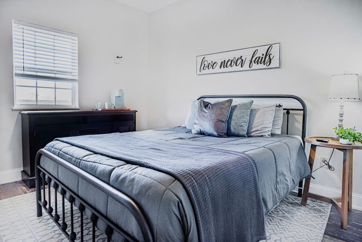 A cozy bedroom featuring a metal bed frame with gray bedding and decorative pillows. A bedside table with a lamp and a small plant is next to the bed, while a dresser and window are visible in the background.