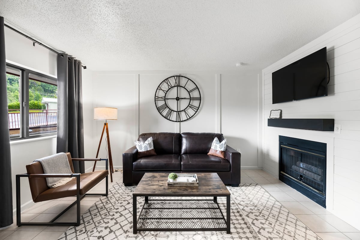 Modern living room featuring a brown leather sofa with decorative pillows, a wooden coffee table, and a wall-mounted television. A floor lamp and large windows with curtains provide natural light, while a wall clock adds a decorative touch.