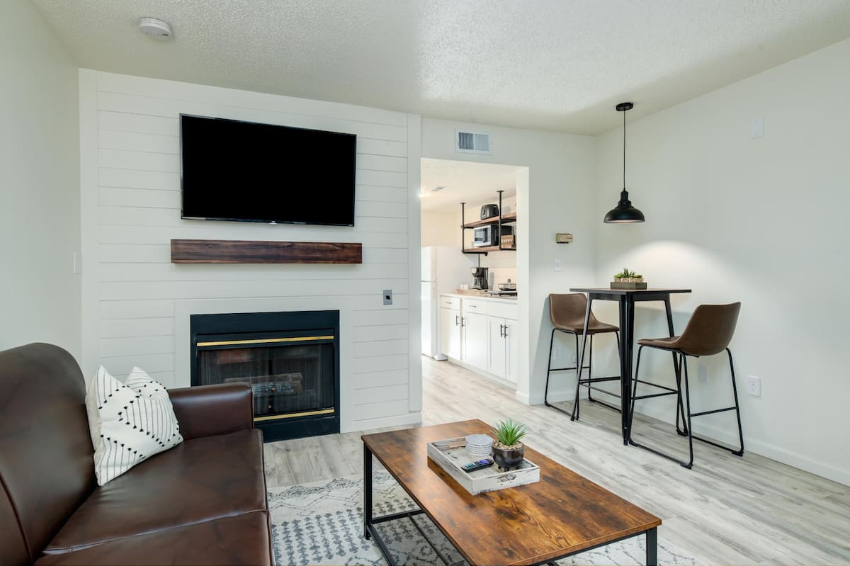 Cozy living area featuring a brown leather sofa, a wooden coffee table, and a mounted flat-screen TV above a fireplace. The background shows a glimpse of the kitchen area with modern appliances and a dining table with two chairs.