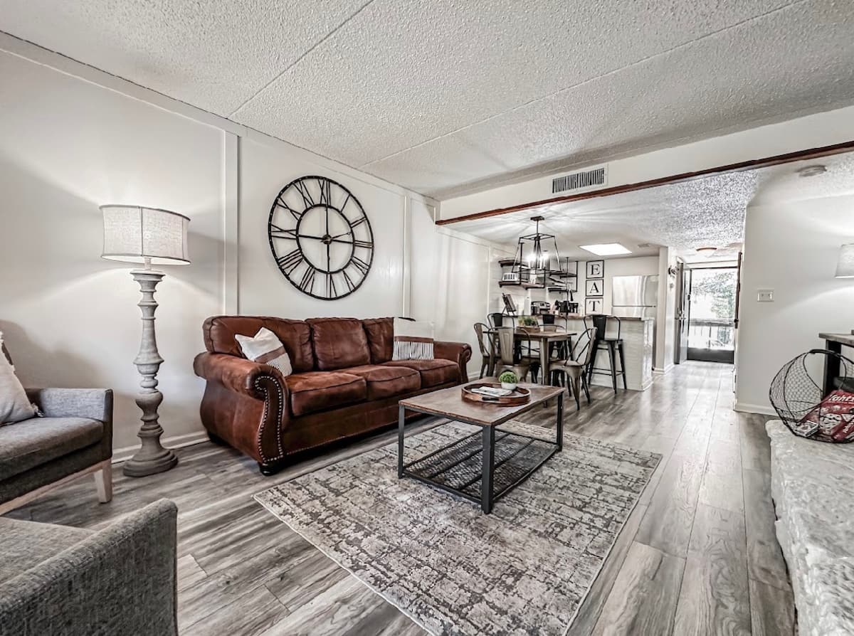Cozy living room with a brown leather sofa, a stylish wooden coffee table, and a decorative rug. A large wall clock is mounted above the couch, and a dining area with black metal chairs is visible in the background.