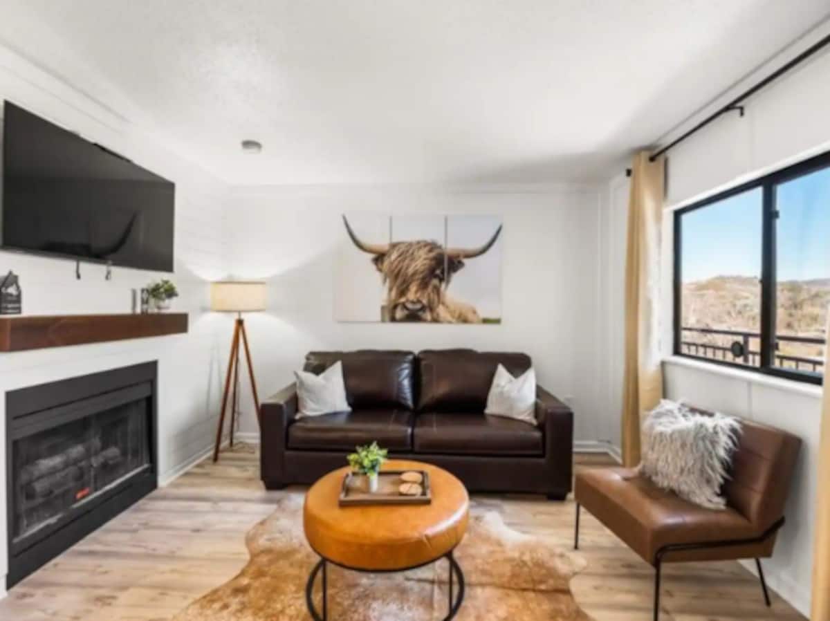 Cozy living room featuring a brown leather sofa, a round coffee table, and a stylish lamp. Artwork of a highland cow is displayed on the wall.