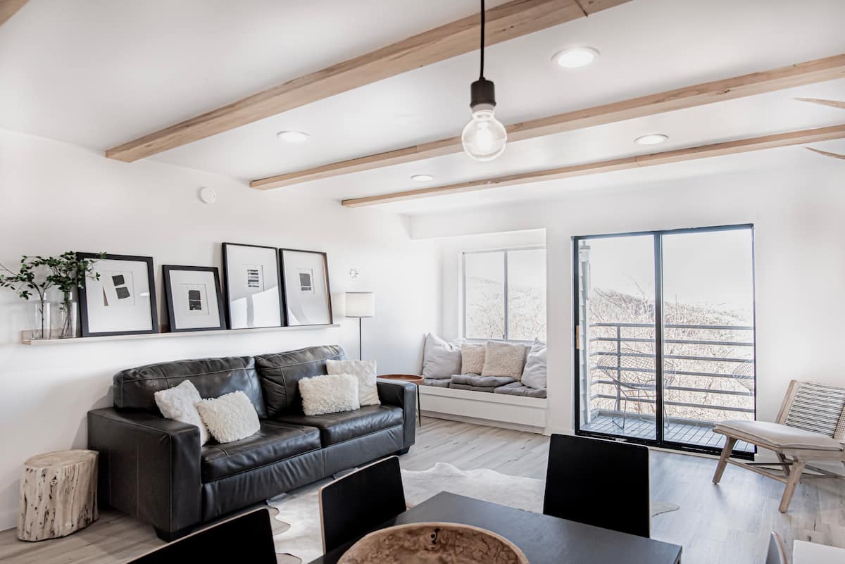 A modern living room with a black leather sofa adorned with throw pillows, wooden beams on the ceiling, and a dining table. Large windows offer scenic views, while a sliding door leads to a balcony.