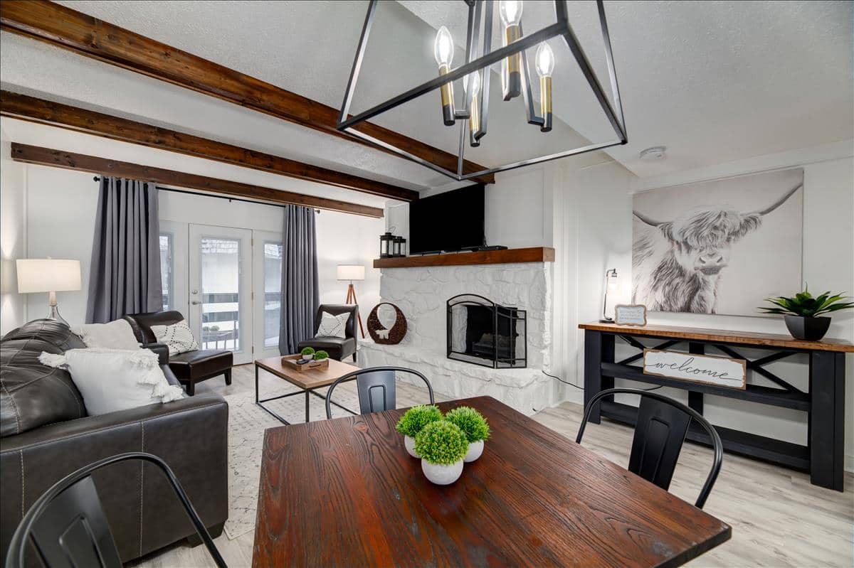 Cozy living room featuring a leather sofa, wooden coffee table, and a dining table with plants, accentuated by exposed beams and a stone fireplace.