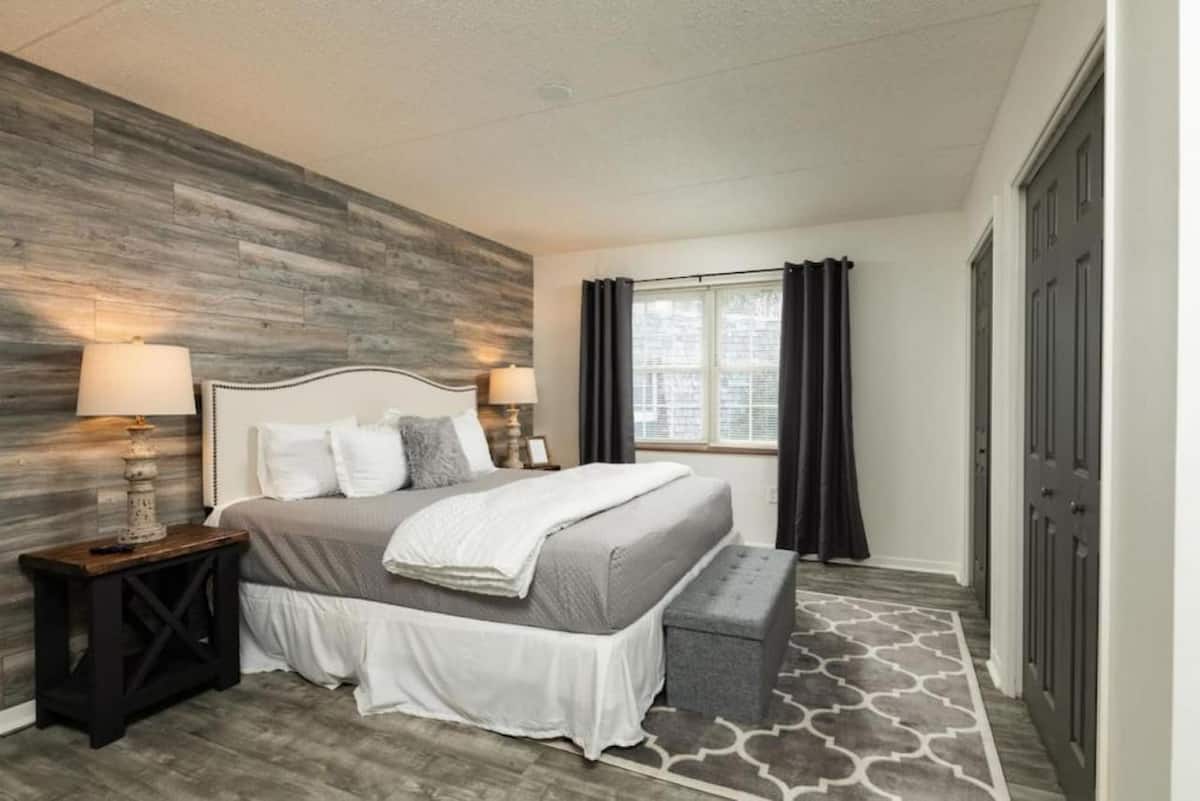 A cozy bedroom featuring a king-sized bed with decorative pillows, a gray and white color scheme, bedside lamps, and a stylish area rug. Large windows with dark curtains allow natural light to brighten the space.