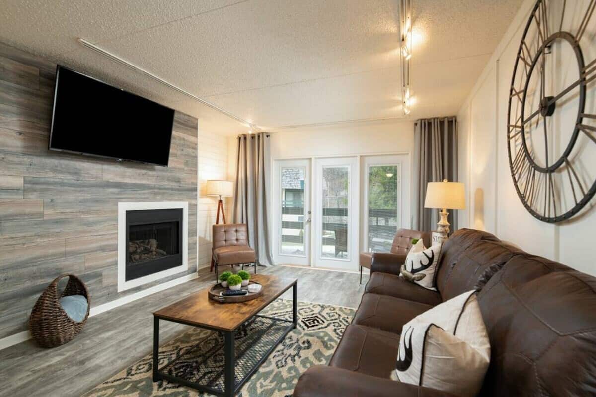 Cozy living room featuring a wall-mounted TV, a modern fireplace, and comfortable brown leather seating, decorated with patterned pillows. A wooden coffee table with a decorative tray sits in the center, while large windows provide natural light.