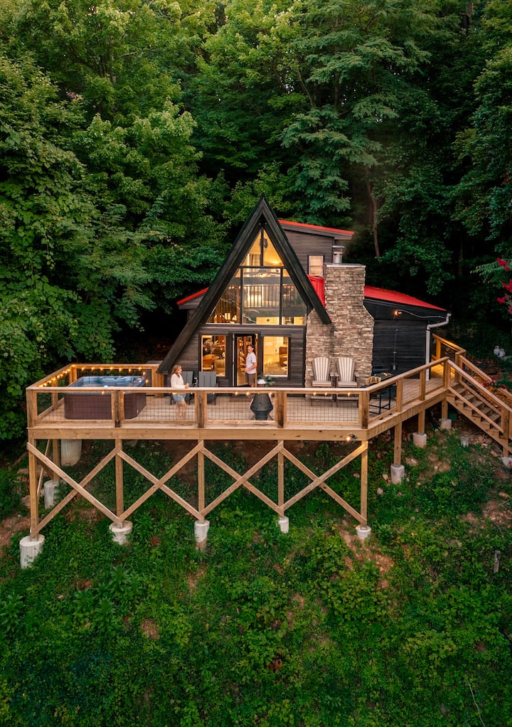 Aerial view of a modern A-frame cabin surrounded by trees, featuring a wooden deck with seating and a hot tub.