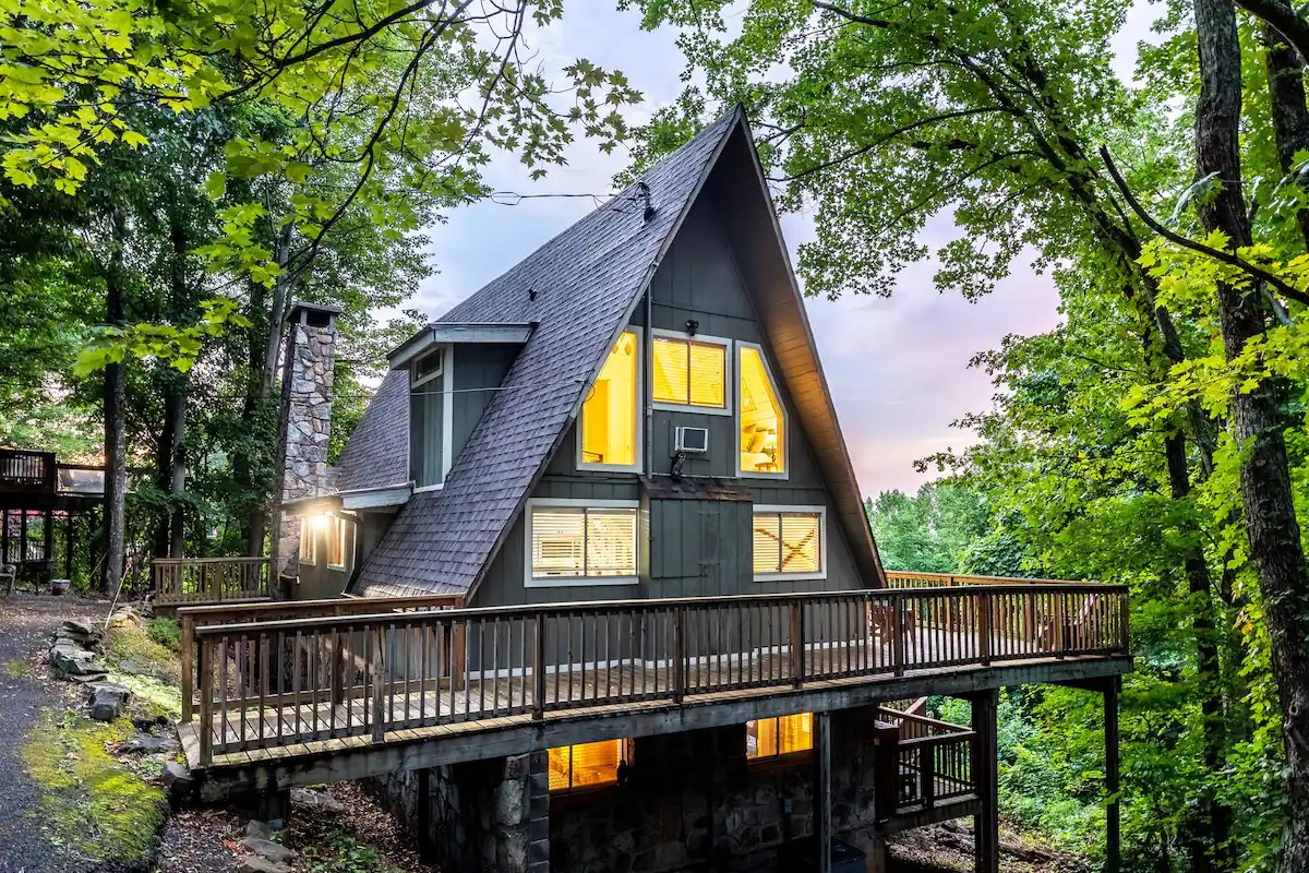 A cozy A-frame cabin surrounded by trees, featuring a wooden deck and large windows glowing with warm light.