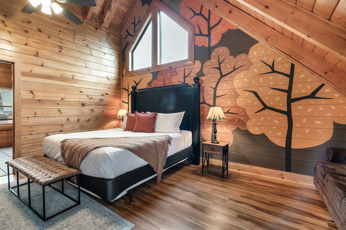 Cozy bedroom in a wooden cabin featuring a large bed with decorative pillows, a bench, and a lamp beside the bed, complemented by a mural of colorful trees on the wall.