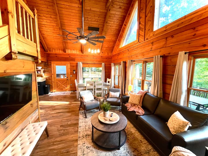 Interior of a cozy cabin featuring wooden walls, high ceilings, comfortable seating, and a dining area with large windows letting in natural light.