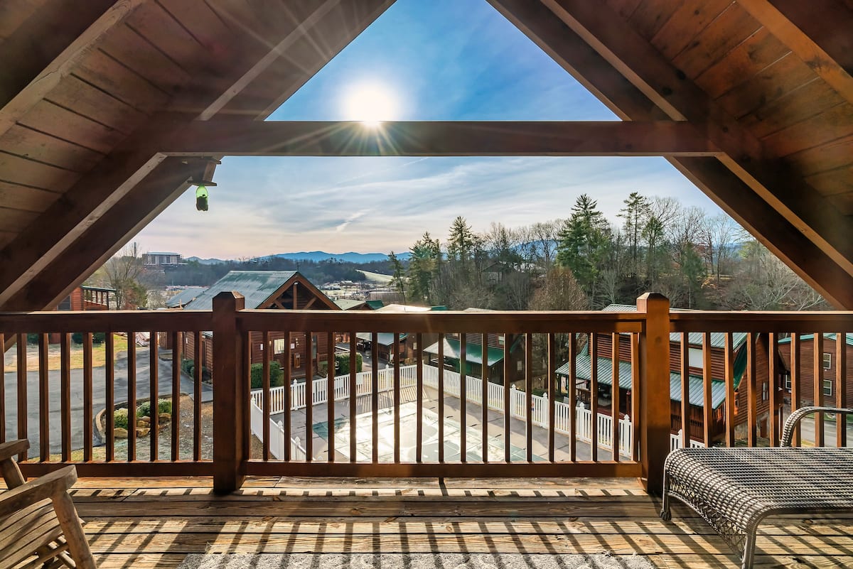 View from a wooden balcony with a railing overlooking a landscape of trees and mountain hills under a bright sky.
