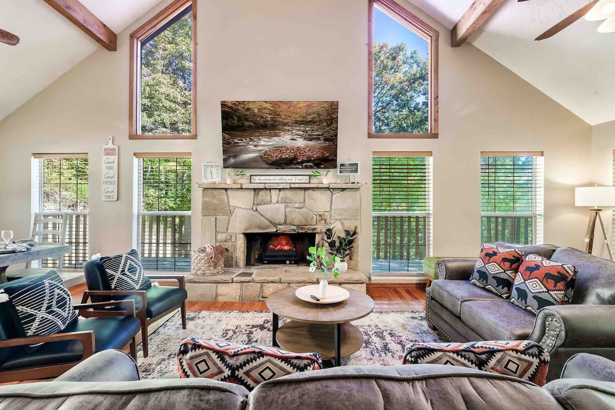 A cozy living room with a stone fireplace, large windows, and comfortable seating. A wall-mounted television displays nature artwork, while a circular coffee table and dining area are visible.