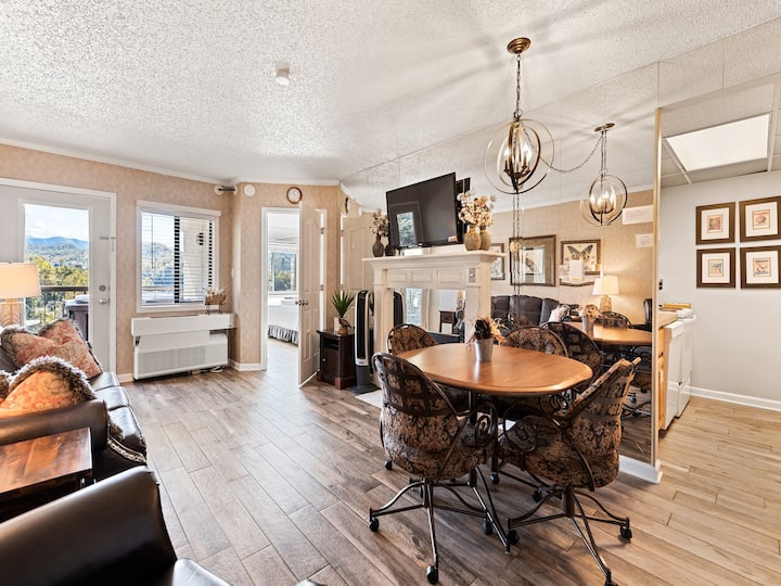 Cozy living area with a sofa and dining table, featuring a mounted television and decorative accents, with a view of mountains through a window.