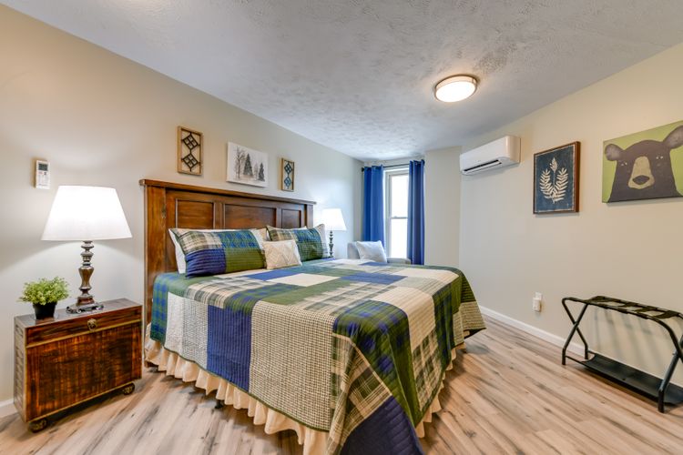 A cozy bedroom featuring a plaid bedspread, bedside lamp, artwork on the walls, and a window with blue curtains.