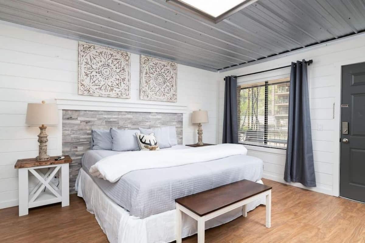 Cozy bedroom featuring a large bed with gray bedding, decorative pillows, and artwork above the headboard. A wooden bedside table with a lamp is next to the bed, and large windows with gray curtains provide natural light.