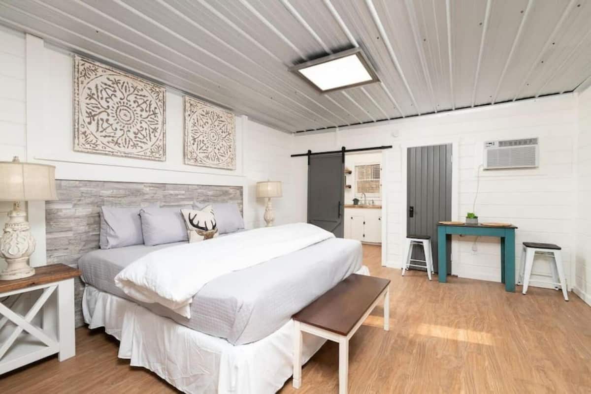 Cozy bedroom with a queen bed, decorative wall art, bedside lamps, and a small table in a bright room with wood flooring.