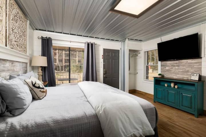 A cozy bedroom featuring a comfortable bed with gray linens, decorative pillows, a bedside lamp, and windows with curtains. There is a television mounted on the wall and a turquoise cabinet nearby. The room has a modern design with a rustic touch.