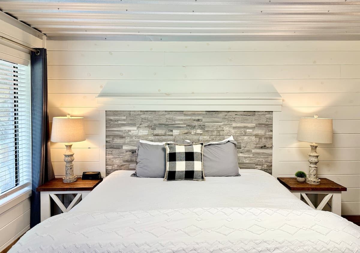 A cozy bedroom featuring a white and gray color scheme, with a wooden headboard, gray pillows, a decorative black and white checkered pillow, bedside lamps, and a wooden side table.