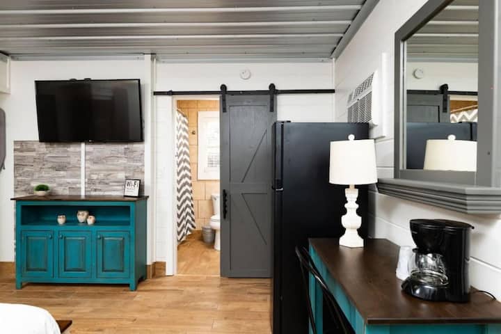 Interior view of a cozy rental unit featuring a mounted flat-screen TV, a teal sideboard, a kitchenette with a refrigerator, a bathroom doorway with a sliding barn door, and a table lamp.