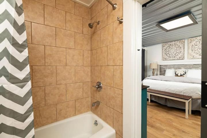Bathroom with a shower and bathtub featuring beige tiles and a chevron shower curtain, leading into a bedroom with a bed and light-colored decor.