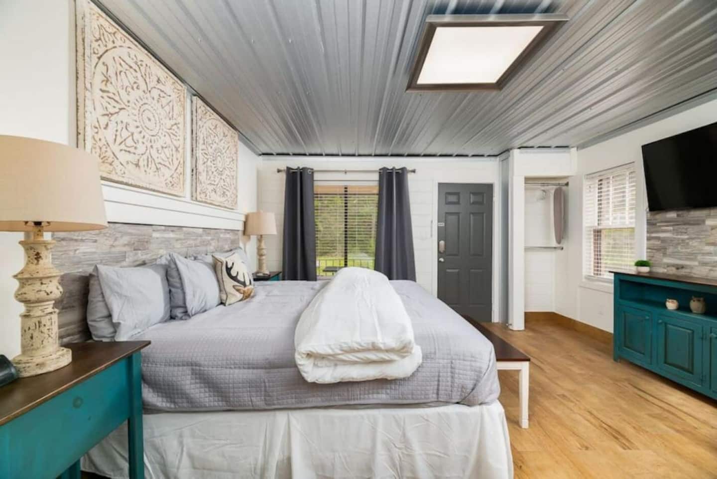 A cozy bedroom featuring a large bed with gray bedding, decorative pillows, a wooden bedside table, and stylish lamps. The room has wooden flooring, a TV mounted on the wall, and a door leading outside. Large windows allow natural light to fill the space.
