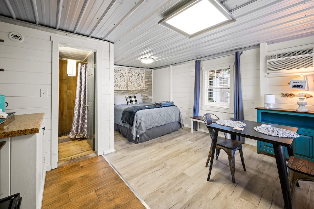 A cozy interior of a small rental space featuring a bed, dining table, and kitchenette, decorated with wooden details and a modern design.