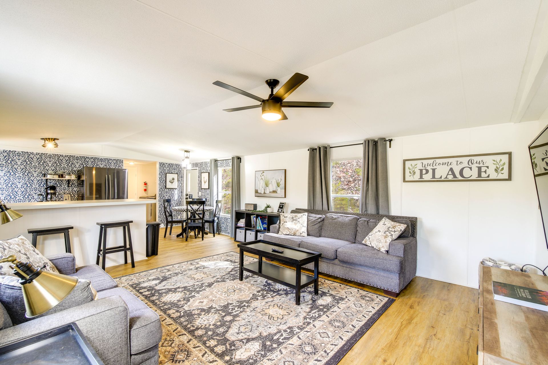 A welcoming living room with a gray sofa, decorative pillows, and a patterned rug, featuring a coffee table and a 'Welcome to Our Place' sign on the wall. The space includes a dining area with black chairs and a modern kitchen in the background.