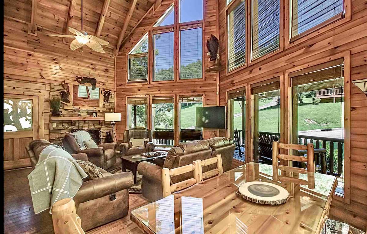 A cozy wooden cabin interior featuring high ceilings, large windows allowing natural light, leather sofas, a wooden dining table, and a stone fireplace.
