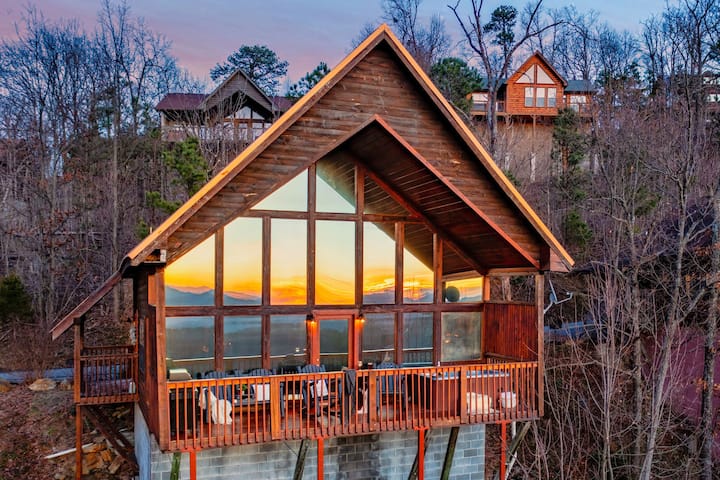 A wooden vacation cabin with large windows, situated on a hillside with sunset views and surrounded by trees.