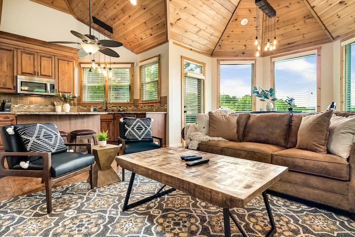 Interior view of a cozy living room featuring a wooden ceiling, a brown sofa, black chairs, and a coffee table, with large windows providing natural light.