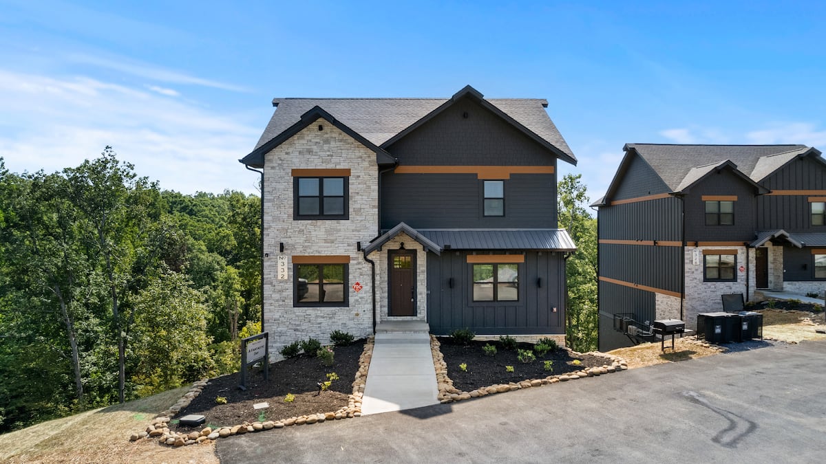 A modern two-story cabin with stone and wood accents, featuring a welcoming front porch and landscaped yard, surrounded by green trees.