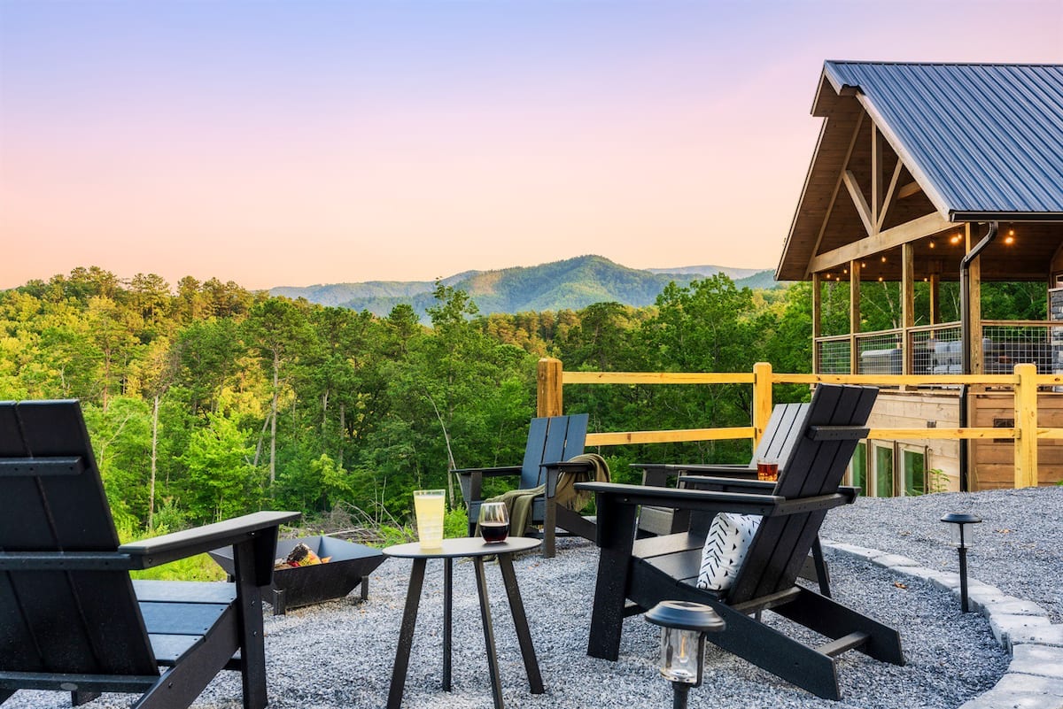 A scenic view from a cabin patio featuring black outdoor chairs and a small table with drinks, surrounded by lush green trees and mountains under a pastel sky.