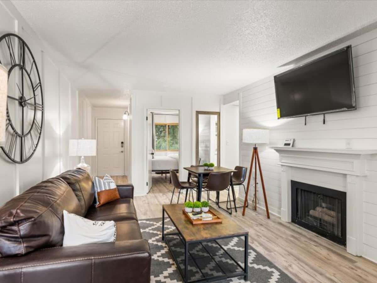 Cozy living area featuring a brown leather sofa, a coffee table with plants, and a large wall clock. There is a mounted TV and a fireplace, with a dining table visible in the background.