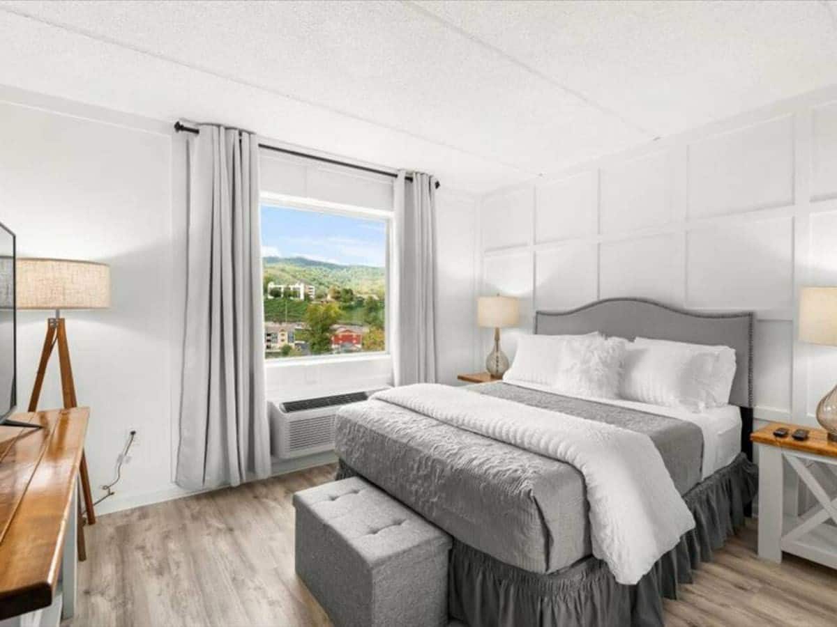 Cozy bedroom with a large window providing a view of the mountains, featuring a queen bed with gray bedding, a nightstand with a lamp, and a television on a wooden stand.