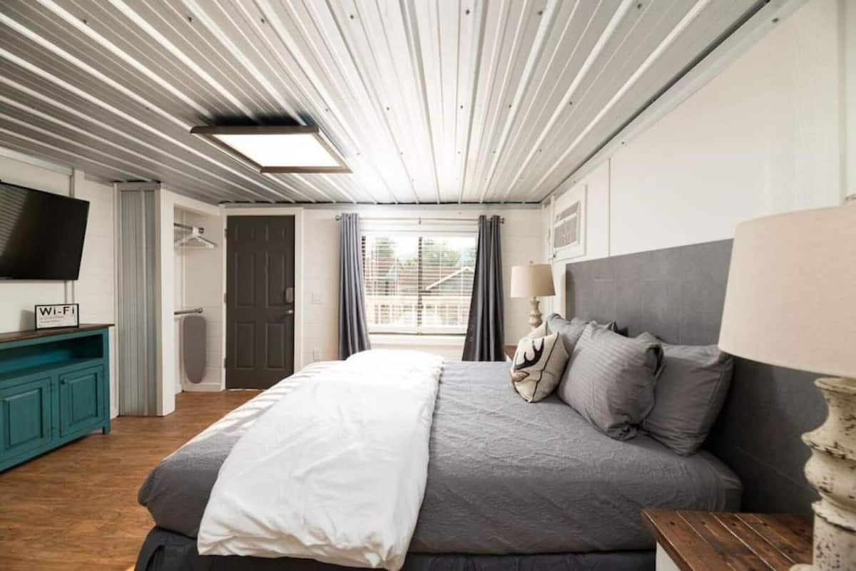 A cozy bedroom featuring a large bed with gray linens, accented with pillows. The room has a rustic design, highlighted by a metallic ceiling, a wooden floor, and a blue cabinet with a television. Natural light streams in through a window with curtains, creating a welcoming atmosphere.