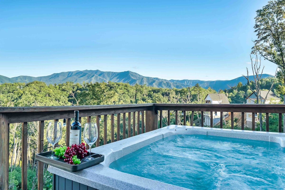 A hot tub on a balcony overlooking green mountains, with a bottle of wine and glasses beside a tray of grapes.