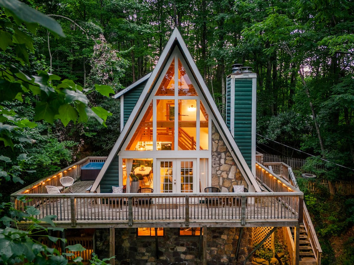 A cozy A-frame cabin surrounded by lush greenery, featuring a wooden deck and large windows showing a warm interior.