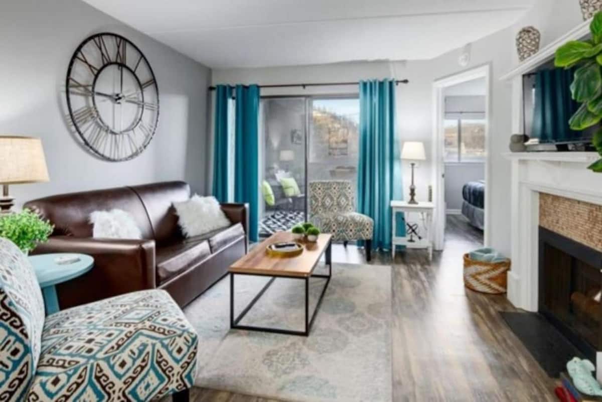 Modern living room featuring a brown leather sofa, decorative armchairs, and a wooden coffee table, with blue curtains and a wall clock.