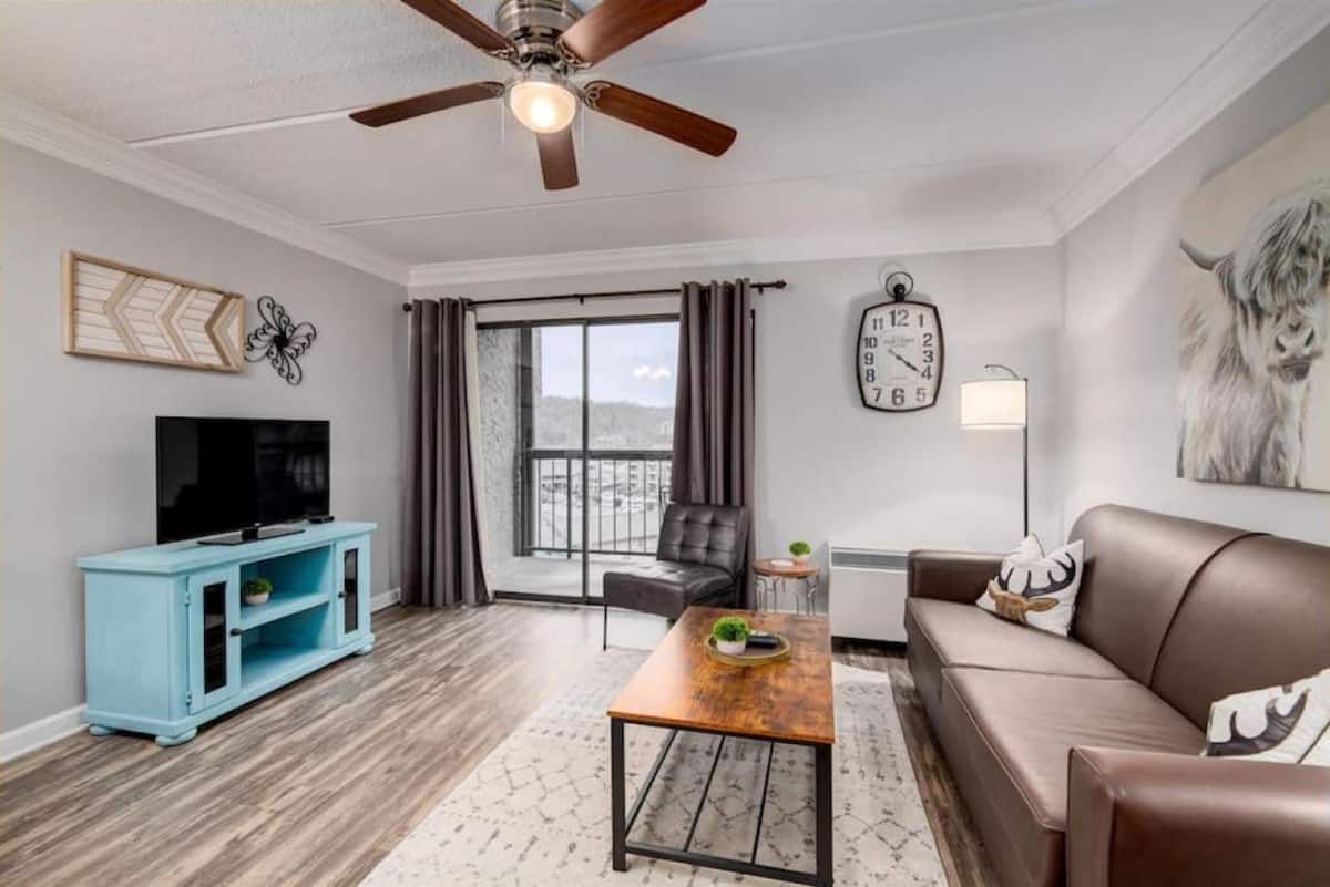A cozy living room featuring a brown leather couch with decorative pillows, a wooden coffee table, and a blue TV cabinet. A large window provides a view of the outdoors, and there is a ceiling fan above.