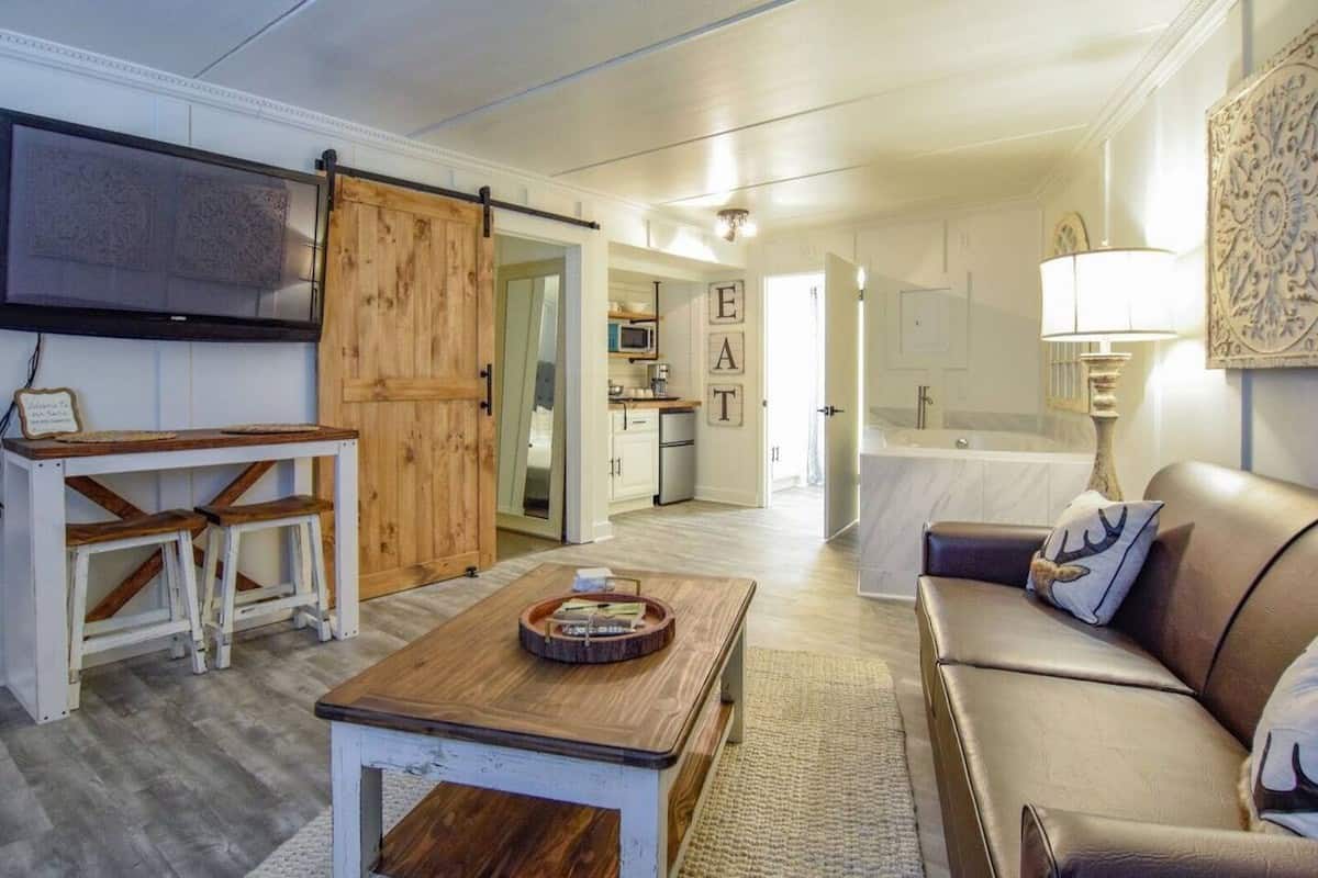 Cozy living area featuring a brown sofa with decorative pillows, a wooden coffee table, and a wall-mounted TV. The space includes a dining area with high stools and a view into a kitchen area with white cabinetry.