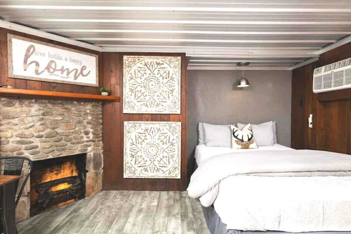 Cozy bedroom featuring a stone fireplace, wooden accents, and decorative panels, with a bed adorned with white linens and a pillow featuring an antler design.