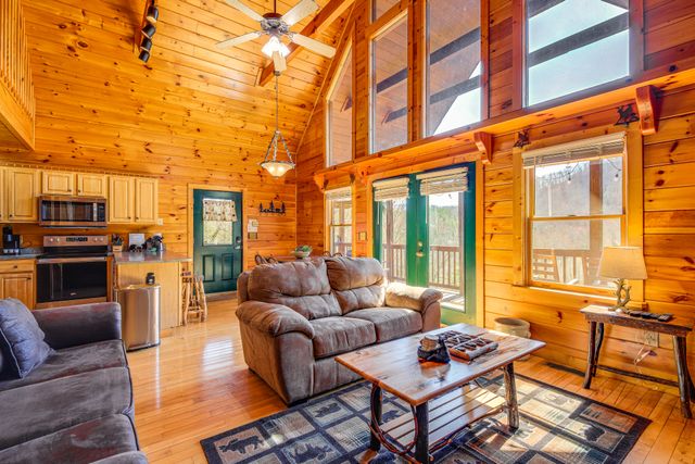 Interior of a cozy log cabin featuring wooden walls, large windows, and comfortable seating. A coffee table and kitchen appliances are visible.
