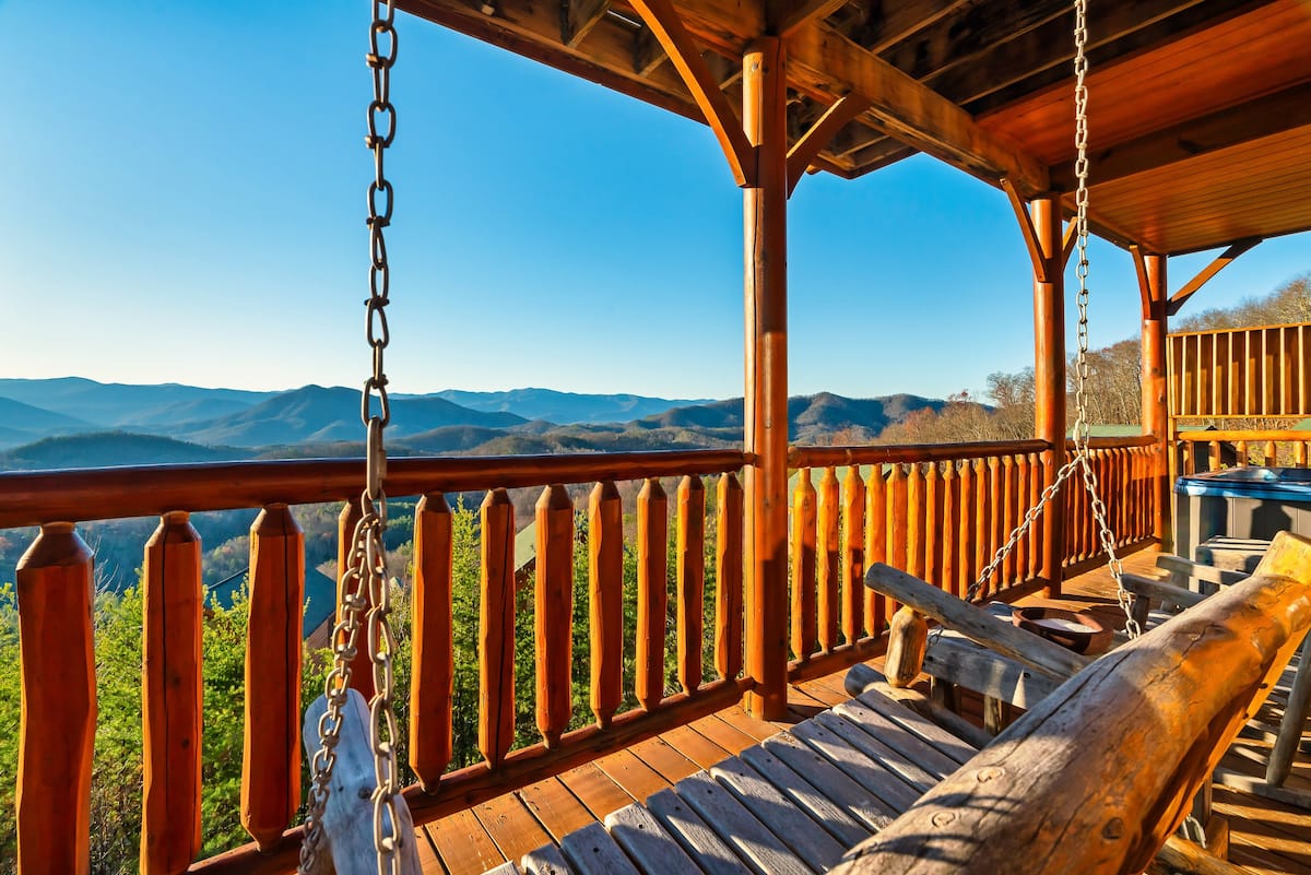 A scenic view from a wooden porch showcasing mountains in the background, complete with swinging chairs and a hot tub.