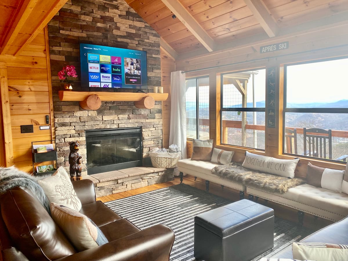 Cozy living room featuring a stone fireplace, plush seating, and a TV mounted on the wall. Large windows offer a scenic view of the outdoors.