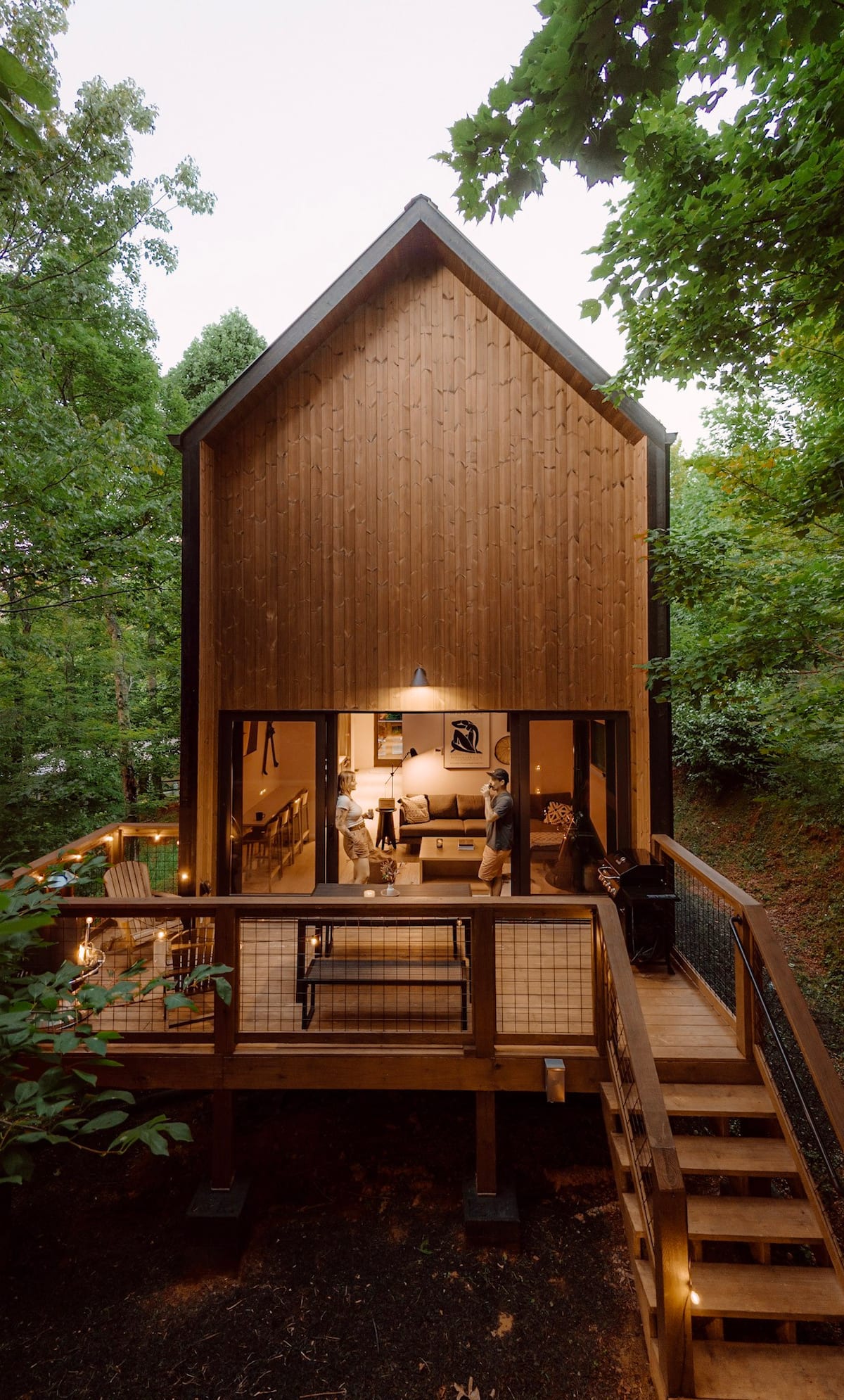 A modern cabin with a wooden exterior surrounded by lush greenery, featuring large glass windows and a deck with outdoor seating. Two individuals are seen inside the cabin enjoying a cozy atmosphere.