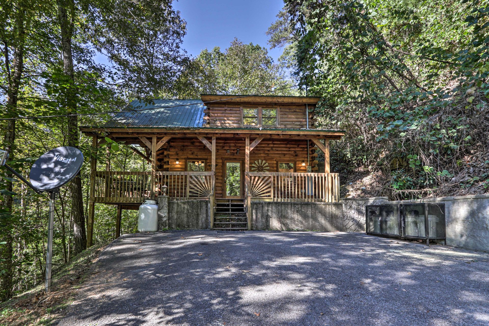 Front view of a wooden cabin surrounded by trees, featuring a porch and a satellite dish.