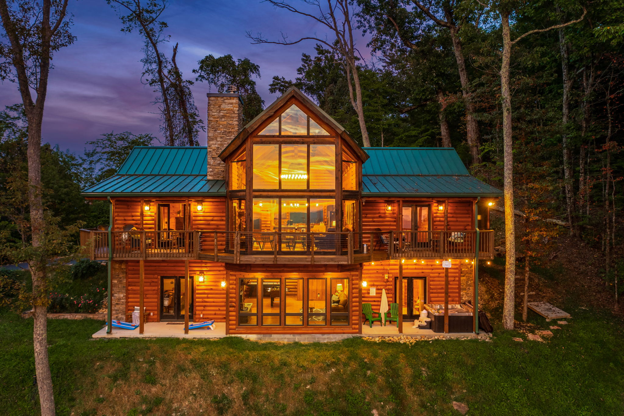 A beautifully lit log cabin surrounded by trees, showcasing large windows and a green roof, with outdoor seating and a hot tub visible.