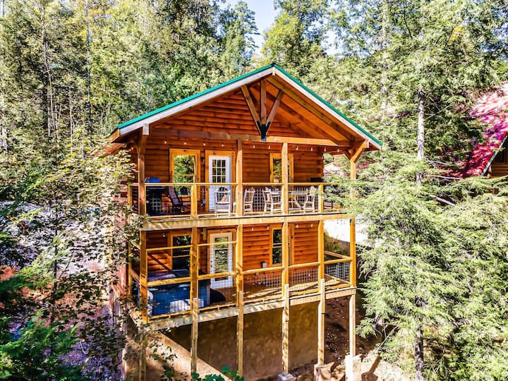 A wooden cabin with a green roof, featuring multiple balconies and surrounded by trees.