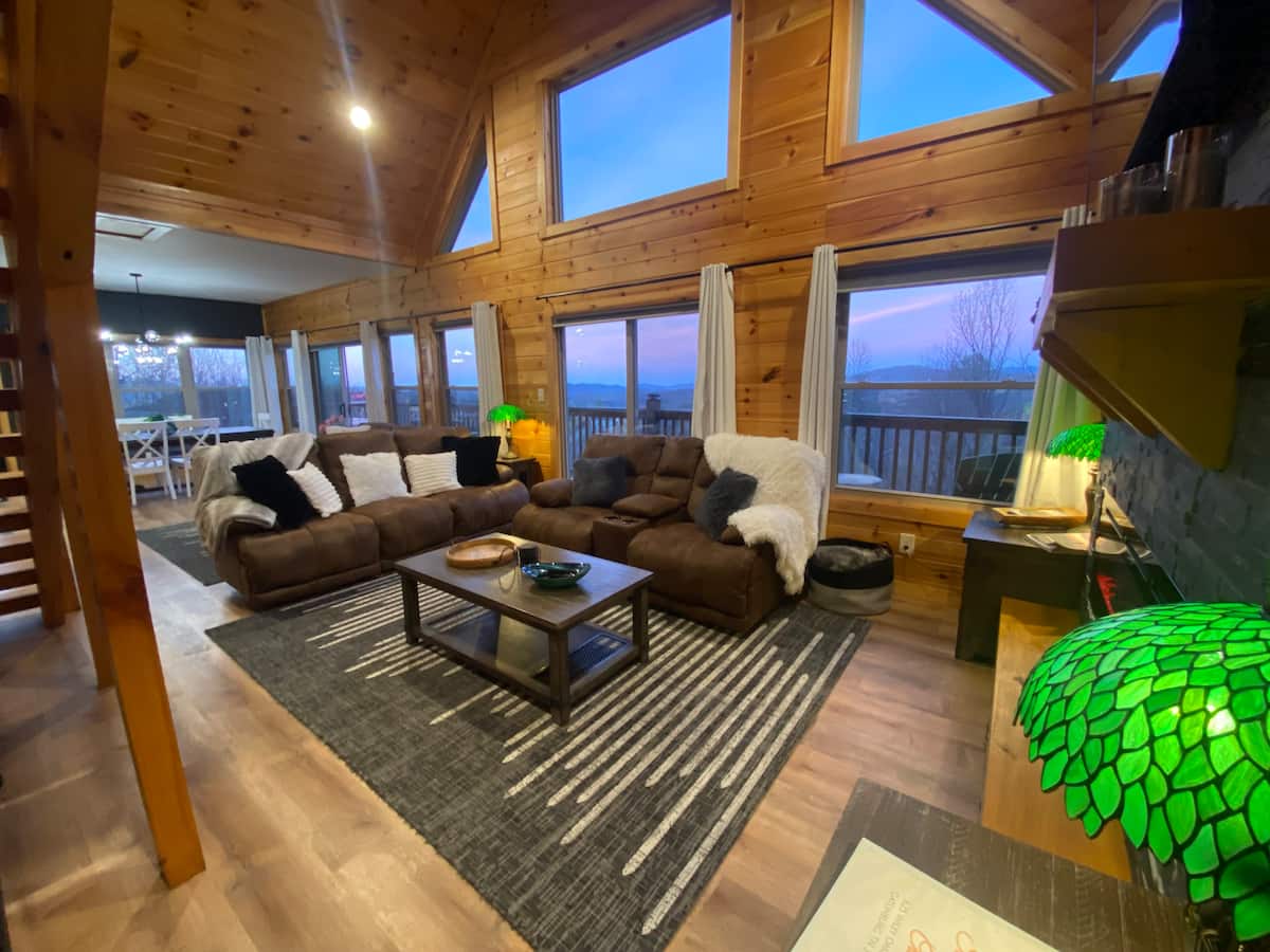 Cozy living room in a wooden cabin with large windows, featuring a brown sectional sofa, a coffee table, and green lamp lights, with scenic views outside.